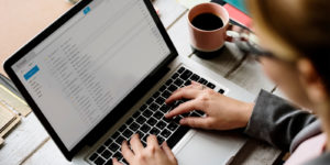 Person typing on silver laptop with pink and brown coffee cup on the desk.