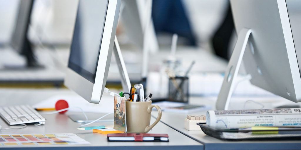 Two computer monitors back to back on desks with various other desk items.