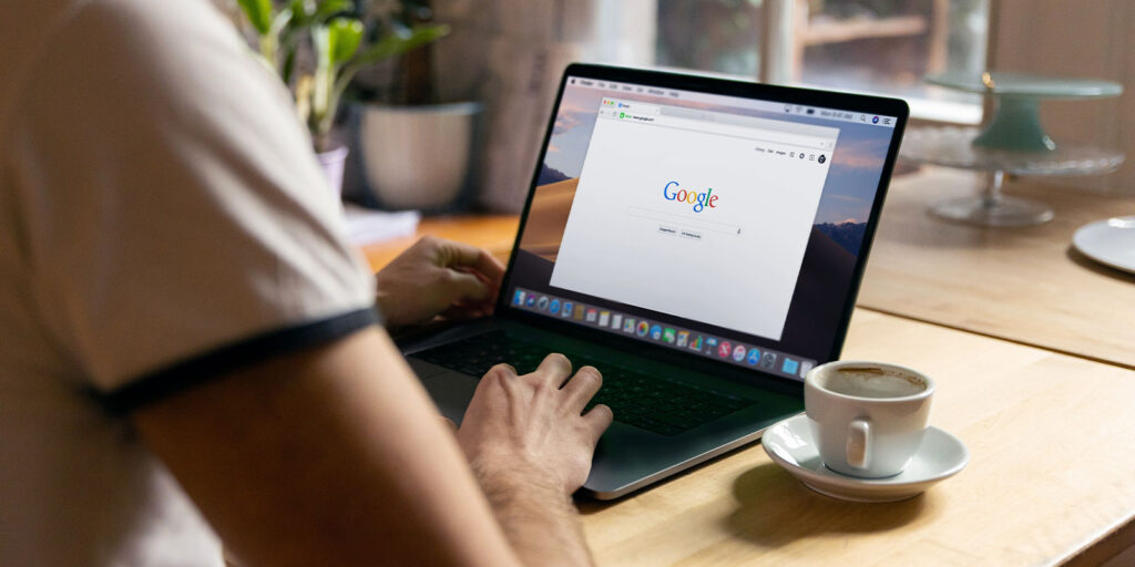 Person typing at a laptop with google open and a white coffee cup on the table.