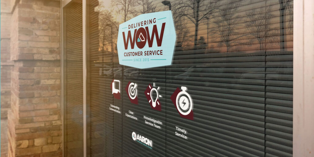 Window with a sign reading “Delivering WOW Customer Service Since 2015” and icons for clean appearance, knowledgeable staff, and timely service. Brick wall and reflections visible—hallmarks of Aaron Overhead Doors’ commitment to excellence.