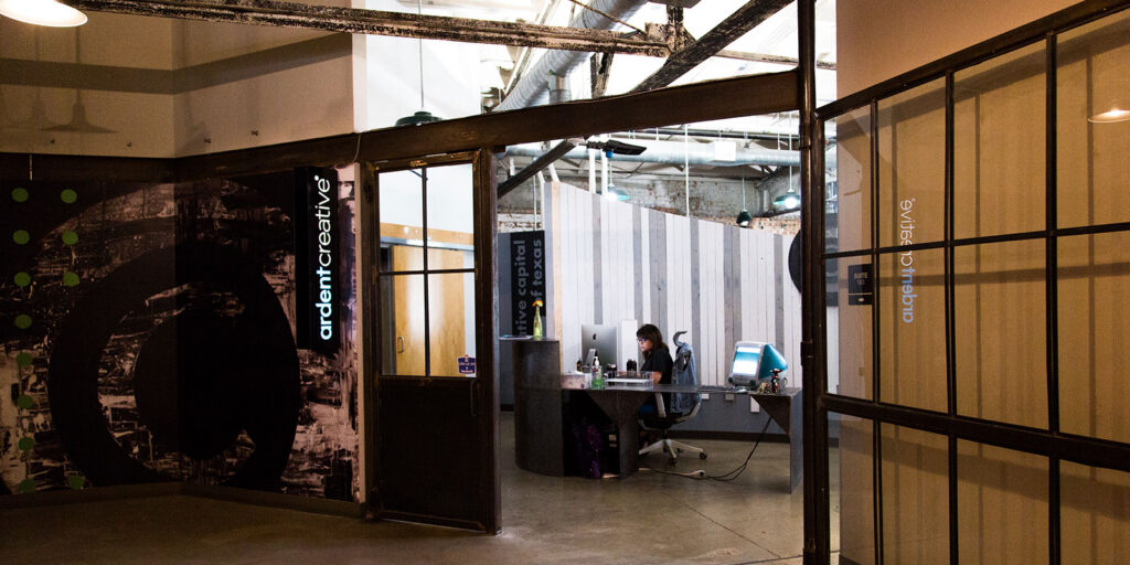 A person works at a computer in a modern office space with glass doors, exposed pipes, and signage reading "Ardent Creative," reflecting the spirit of logo design and brand evolution.
