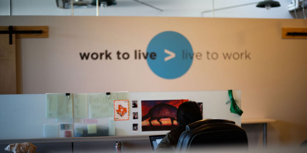 Office space with a person working at a desk; wall behind displays the phrase "work to live > live to work" in large text, reflecting an ardent approach to brand evolution and inspiring thoughtful logo design.