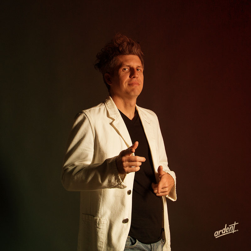 A man in a white blazer and black shirt stands against a dark background, pointing forward with both hands. The word "ardent" appears in the bottom right corner.
